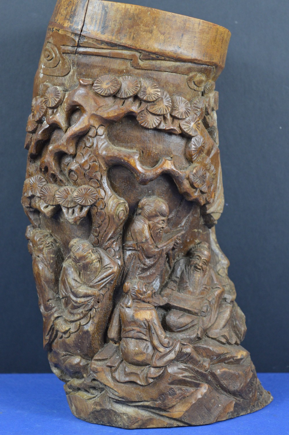 A hand-carved cylindrical bamboo brush pot featuring figures and pine trees carved in high relief against a warm brown wooden surface.