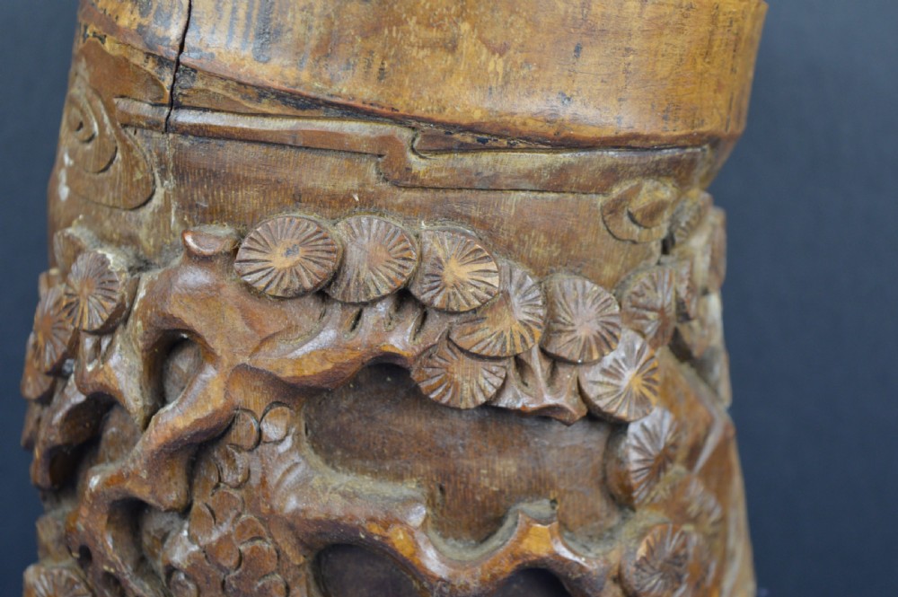 A hand-carved cylindrical bamboo brush pot featuring figures and pine trees carved in high relief against a warm brown wooden surface. View III