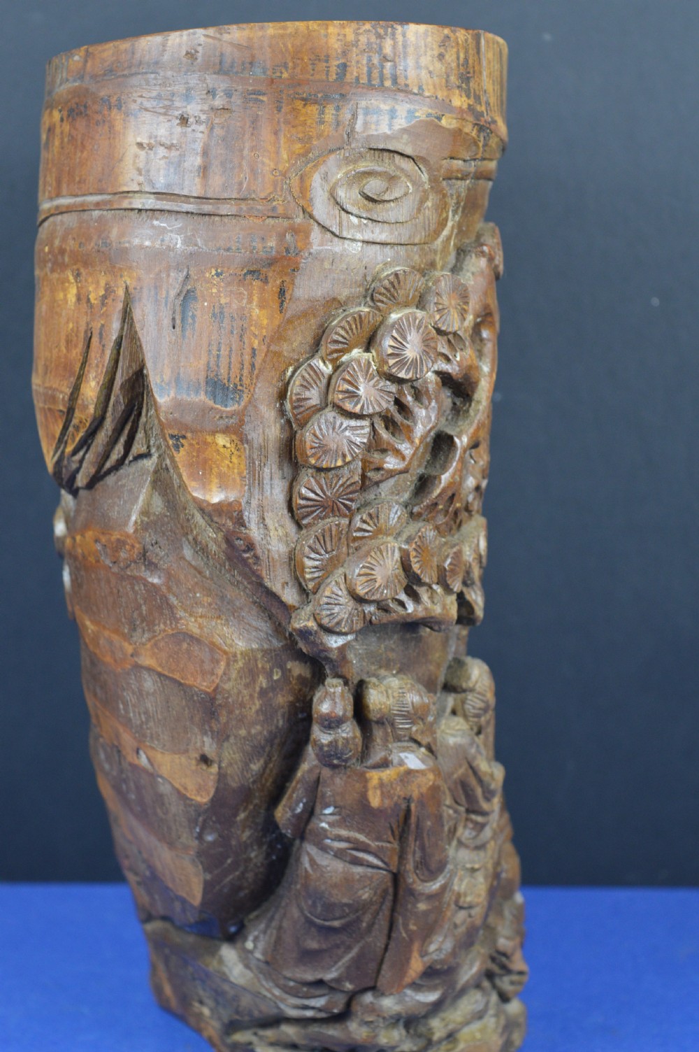 A hand-carved cylindrical bamboo brush pot featuring figures and pine trees carved in high relief against a warm brown wooden surface. View IV