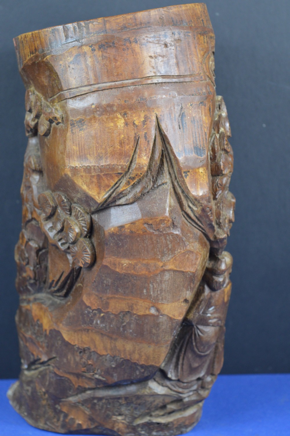 A hand-carved cylindrical bamboo brush pot featuring figures and pine trees carved in high relief against a warm brown wooden surface. View V
