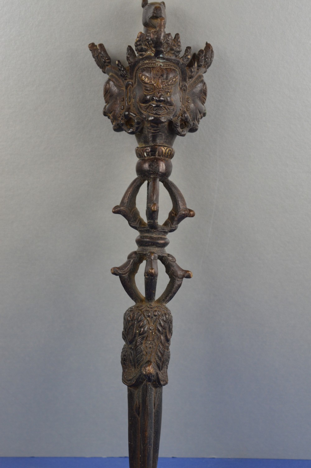 A 19th-century Tibetan bronze phurba featuring a wrathful deity head pommel, central vajra handles, and a three-sided blade extending from a makara mouth.