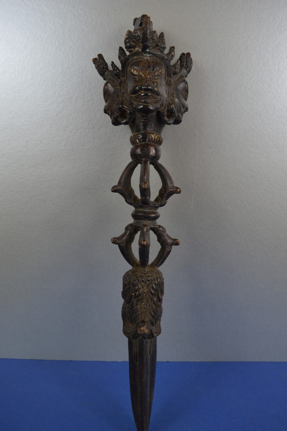 A 19th-century Tibetan bronze phurba featuring a wrathful deity head pommel, central vajra handles, and a three-sided blade extending from a makara mouth. View V