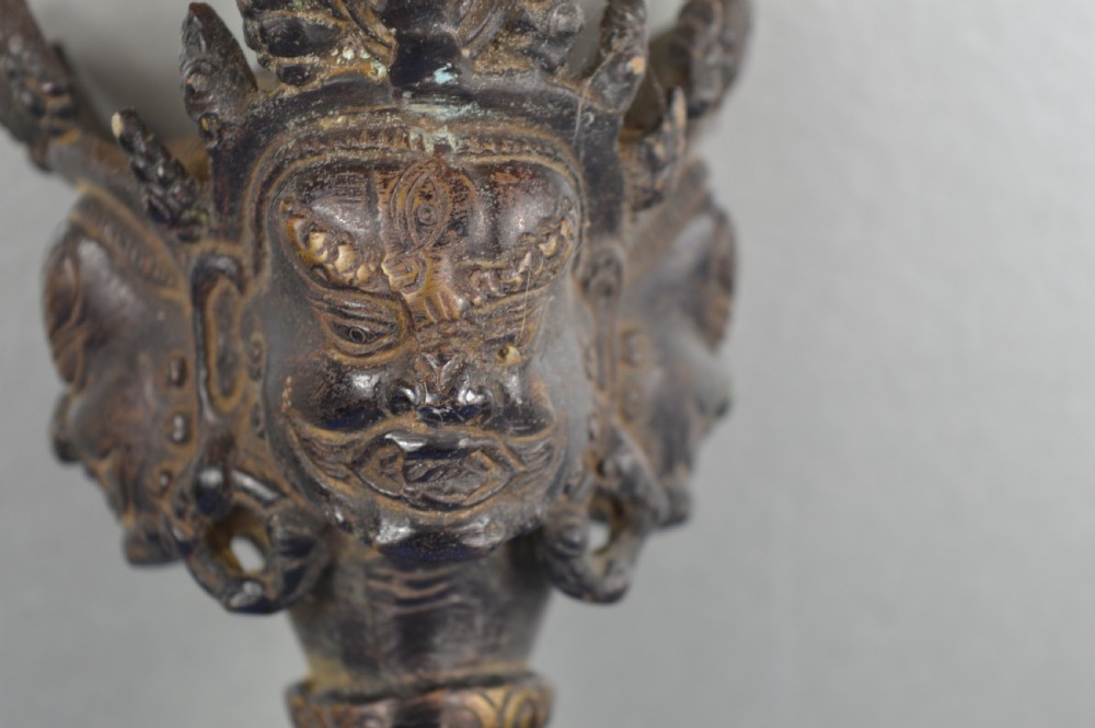 A 19th-century Tibetan bronze phurba featuring a wrathful deity head pommel, central vajra handles, and a three-sided blade extending from a makara mouth. View VI