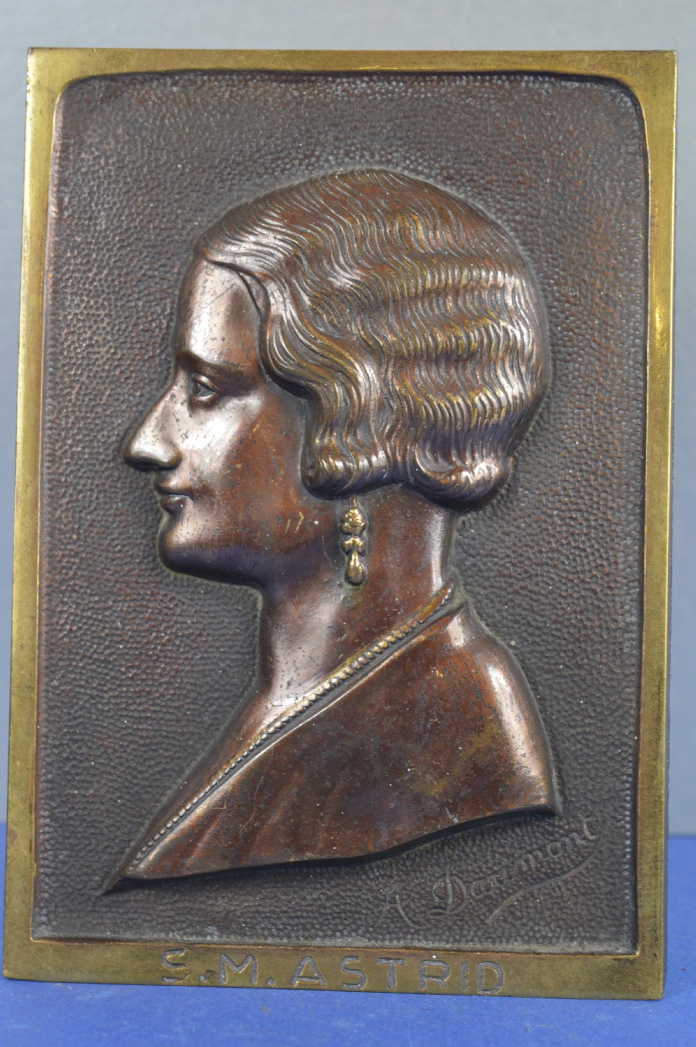 An Art Deco dark brown bronze relief plaque featuring a side profile portrait of Queen Astrid against a textured background with inscribed lettering.