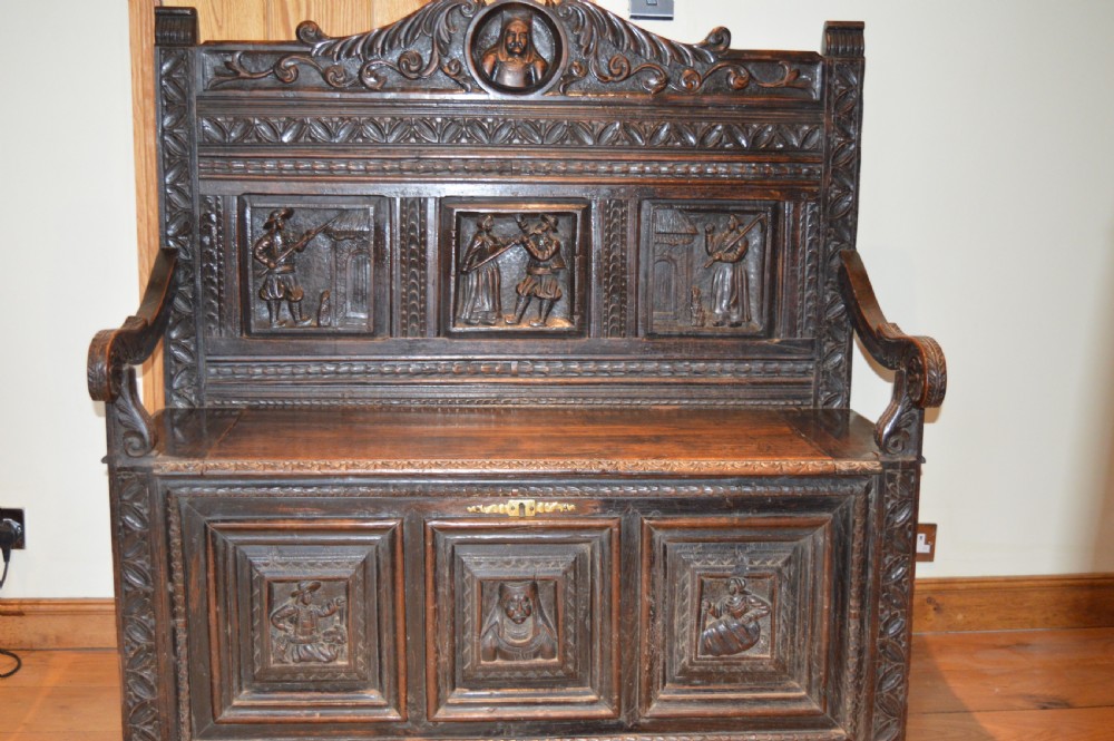 An 18th-century Flemish dark carved oak settle featuring figurative relief panels on the backrest and chest base with scrolled armrests.
