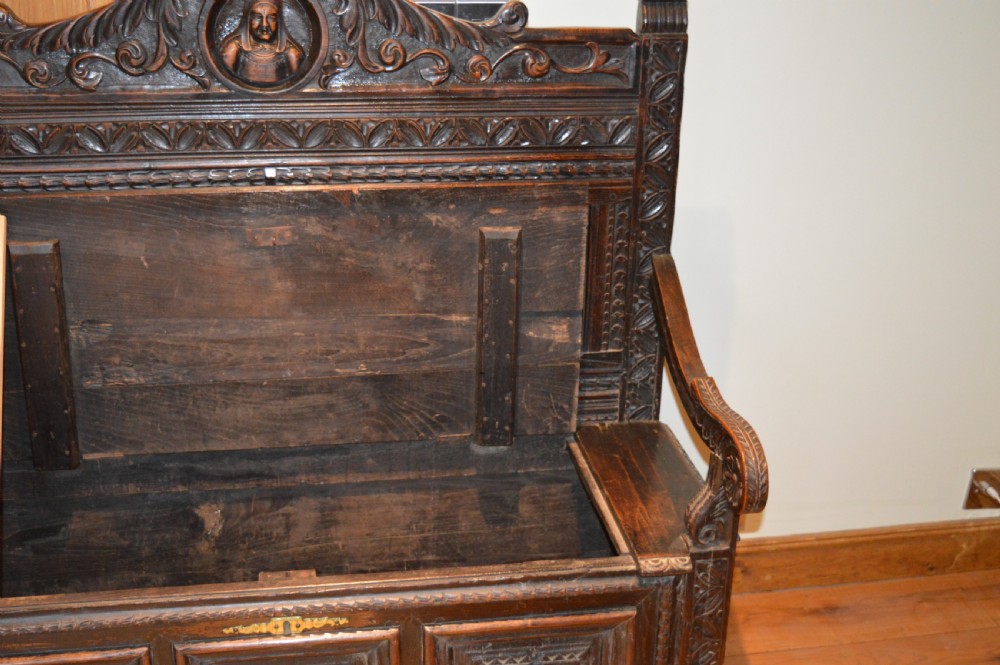 An 18th-century Flemish dark carved oak settle featuring figurative relief panels on the backrest and chest base with scrolled armrests. View XIV
