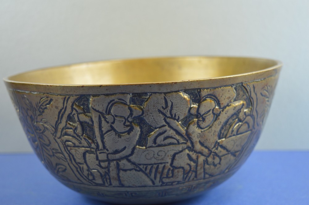 A large circular bronze bowl with relief engravings of figures in a landscape around the exterior and a polished interior.