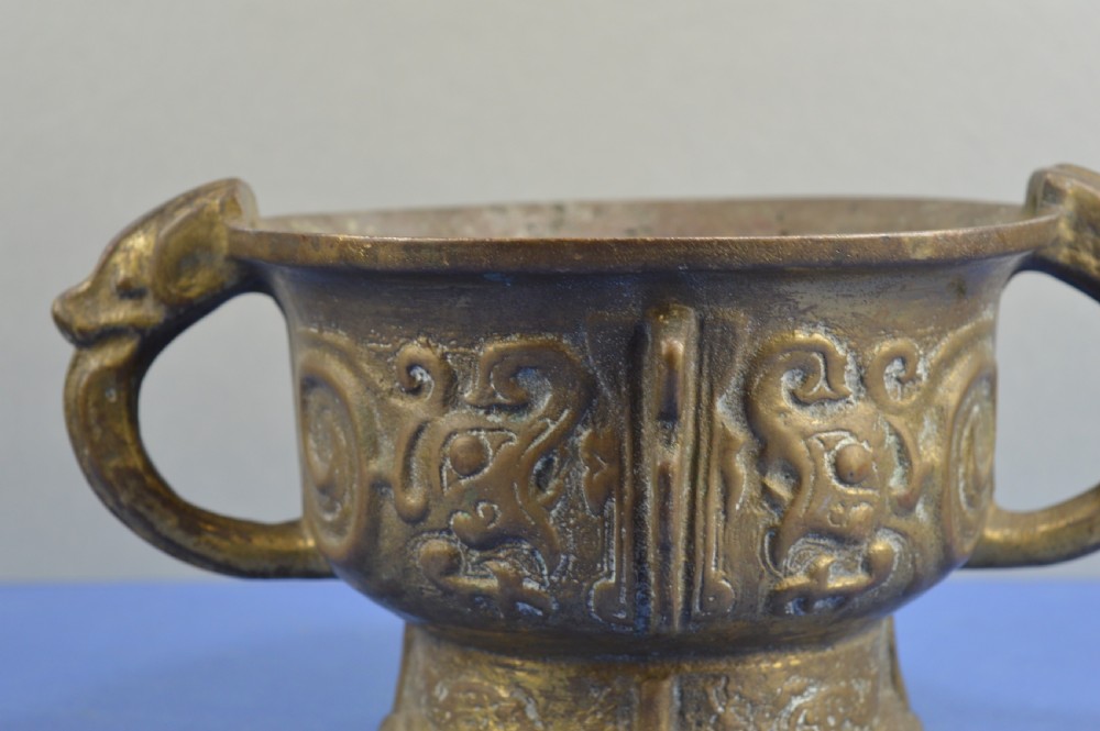 A cast bronze ritual food vessel with two loop handles topped with beast heads, featuring relief carvings and a raised circular base. View IV