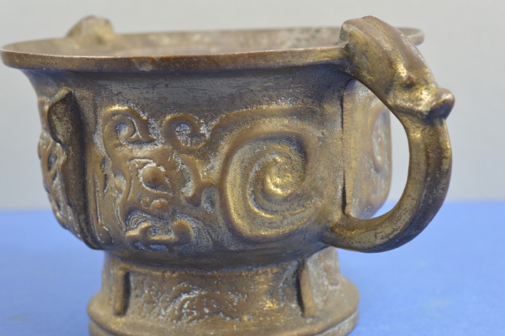 A cast bronze ritual food vessel with two loop handles topped with beast heads, featuring relief carvings and a raised circular base. View V