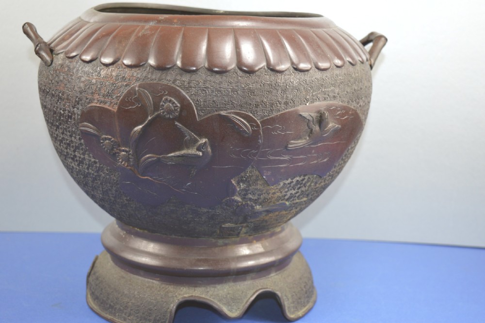 A heavy dark bronze Chinese bowl featuring relief panels with bird and floral motifs, bamboo-style side handles, and a lobed collar over an integral stepped base.