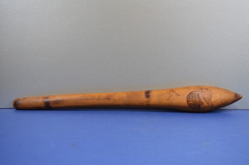 A carved light brown hardwood throwing club featuring a pointed bulbous head with pyrography markings and a smooth tapered shaft. View II