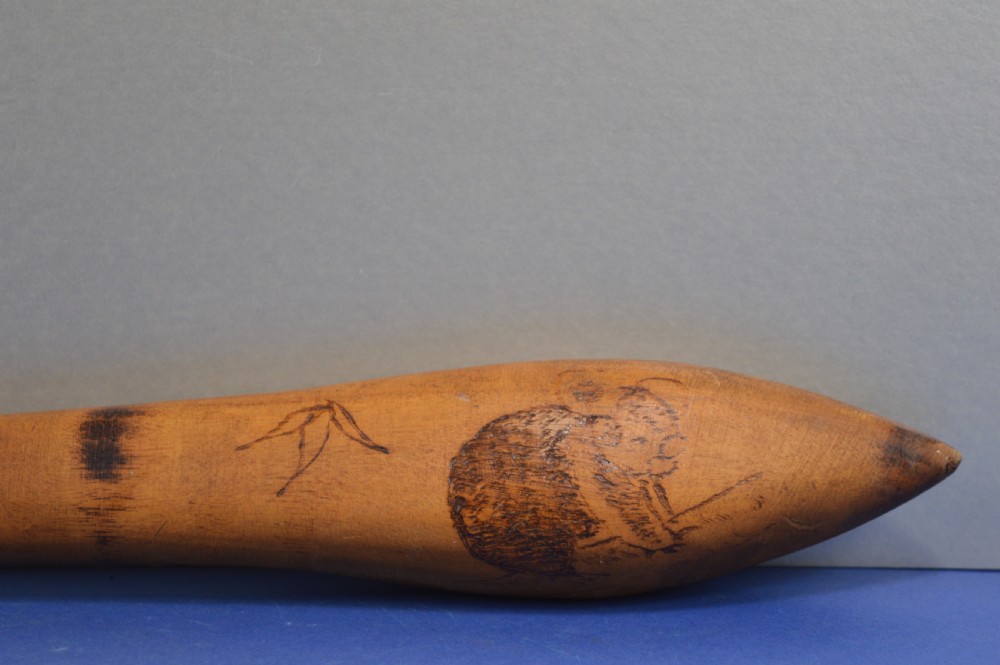 A carved light brown hardwood throwing club featuring a pointed bulbous head with pyrography markings and a smooth tapered shaft. View III