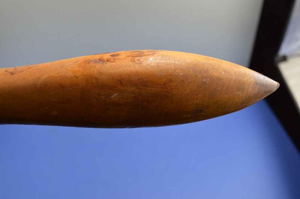 A carved light brown hardwood throwing club featuring a pointed bulbous head with pyrography markings and a smooth tapered shaft. View XIV