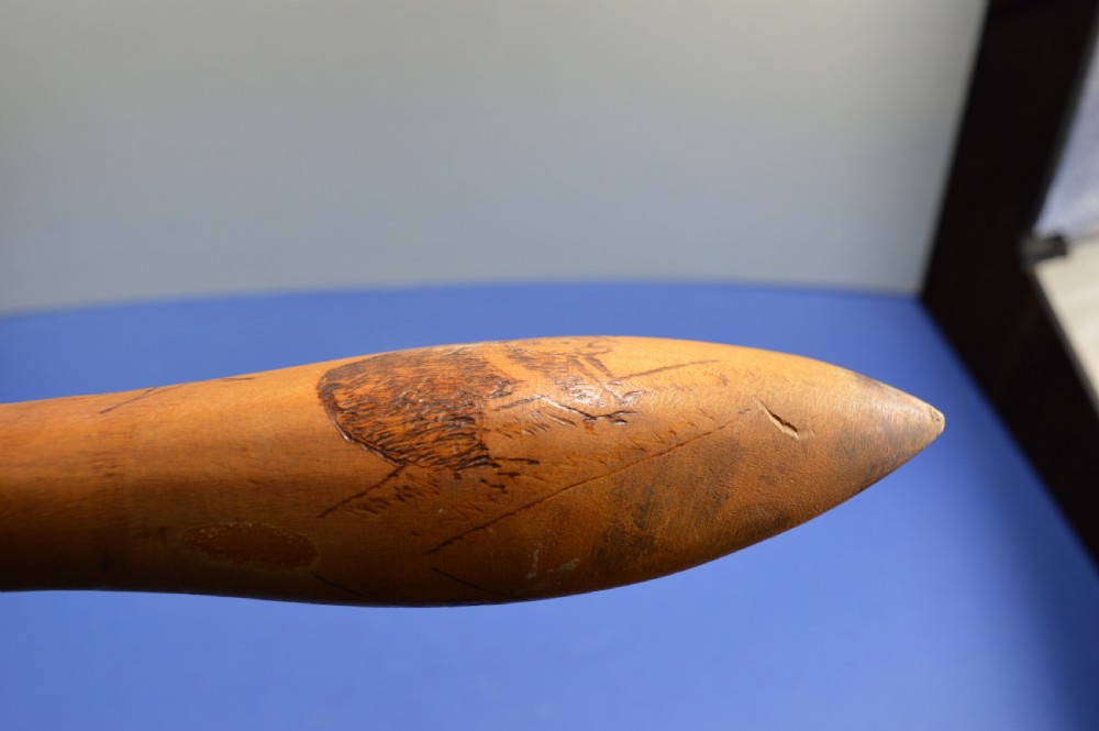 A carved light brown hardwood throwing club featuring a pointed bulbous head with pyrography markings and a smooth tapered shaft. View XV