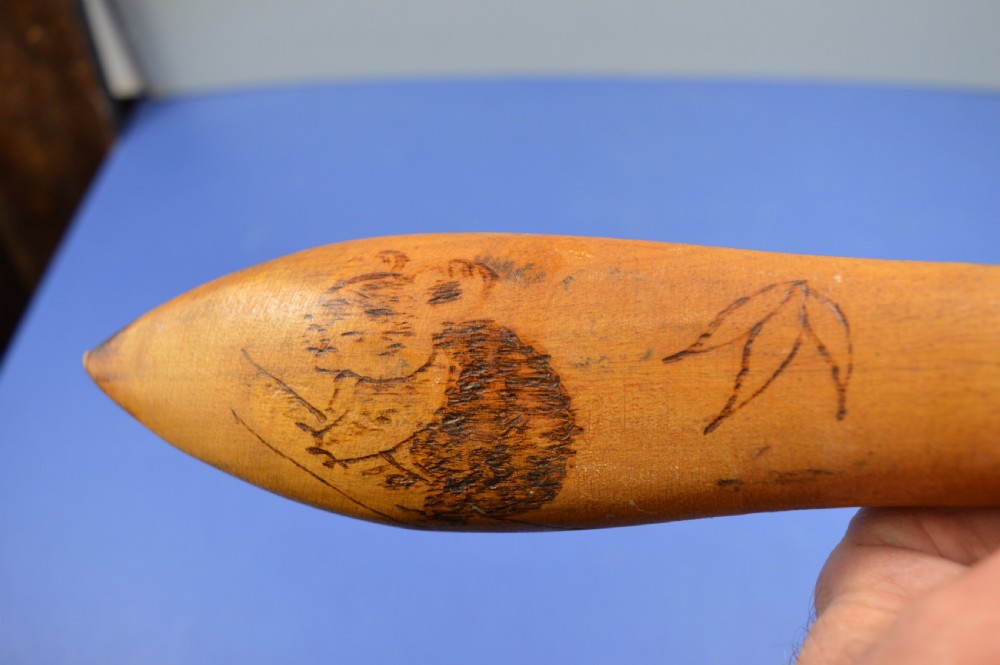 A carved light brown hardwood throwing club featuring a pointed bulbous head with pyrography markings and a smooth tapered shaft. View XVI