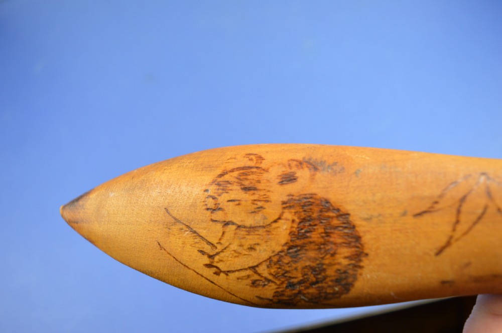 A carved light brown hardwood throwing club featuring a pointed bulbous head with pyrography markings and a smooth tapered shaft. View XVII