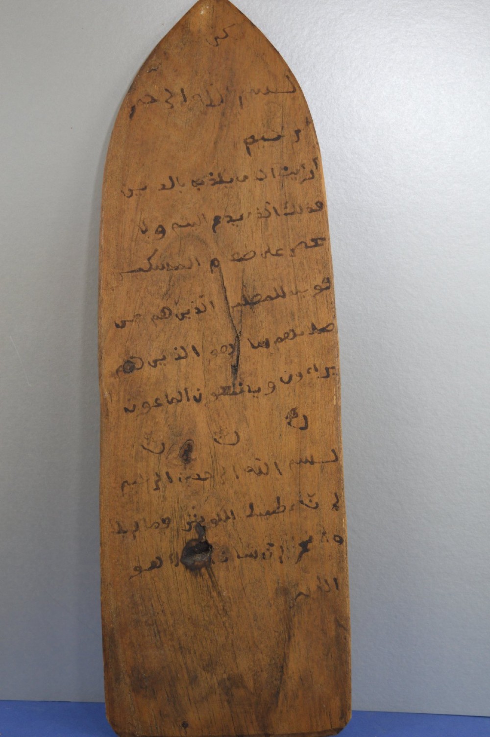 An elongated wooden tablet featuring handwritten Arabic calligraphy in black ink on a natural wood surface.