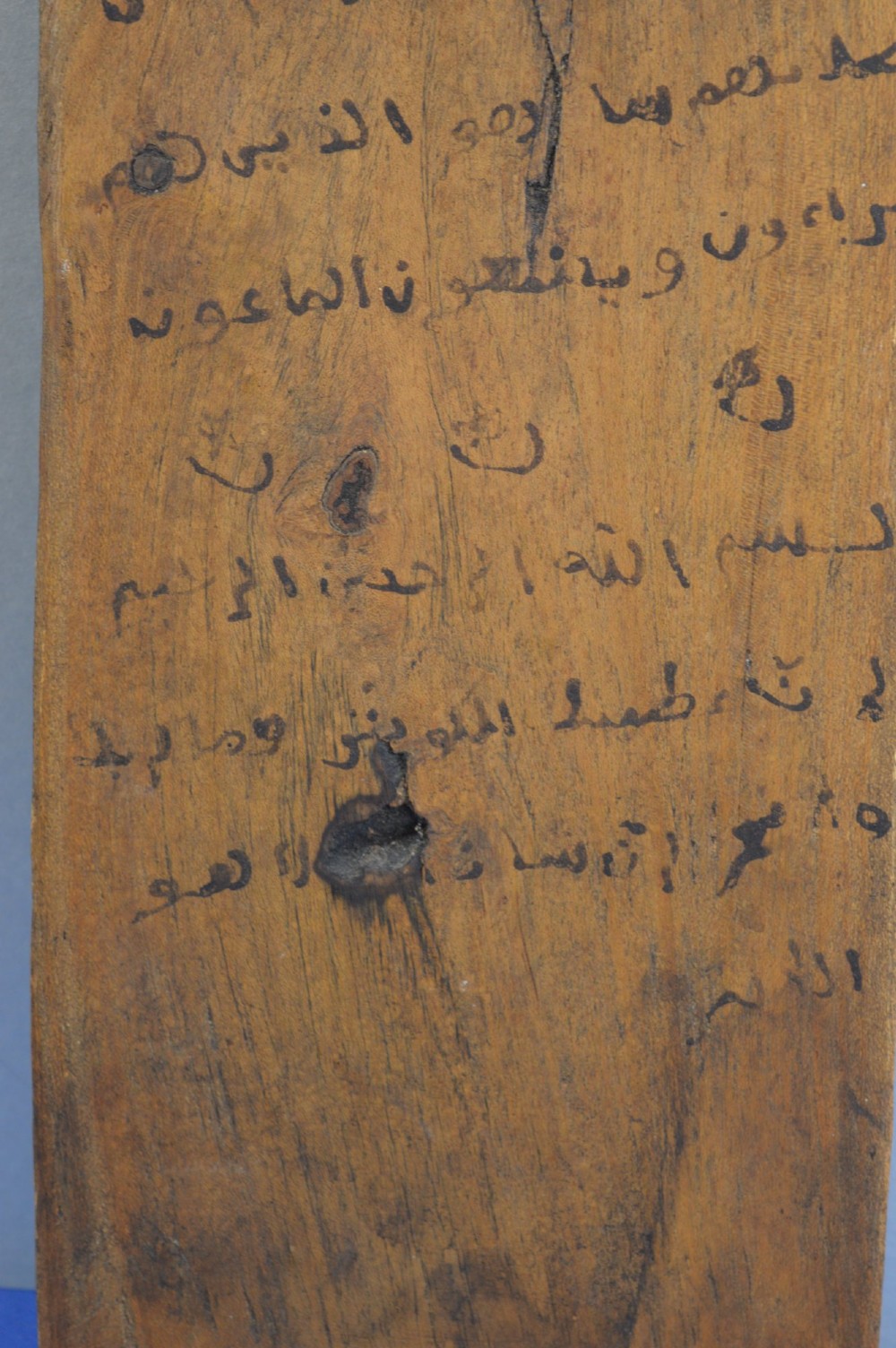 An elongated wooden tablet featuring handwritten Arabic calligraphy in black ink on a natural wood surface. View III