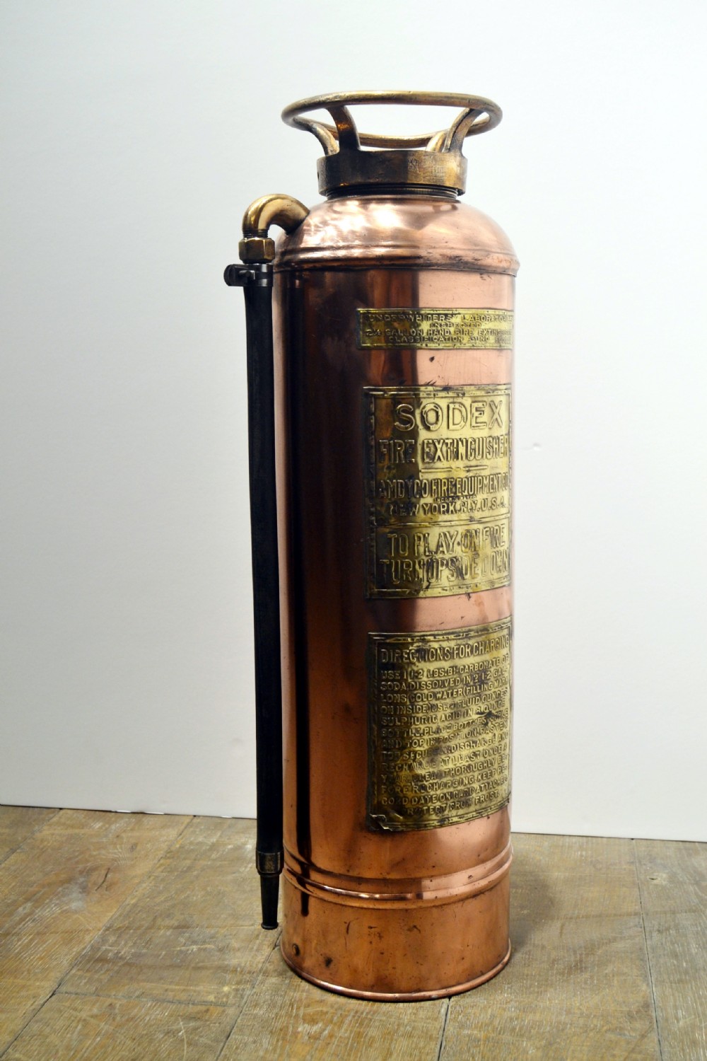 A cylindrical antique copper fire extinguisher featuring brass handles, brass instruction plaques, and a side-mounted black rubber hose.