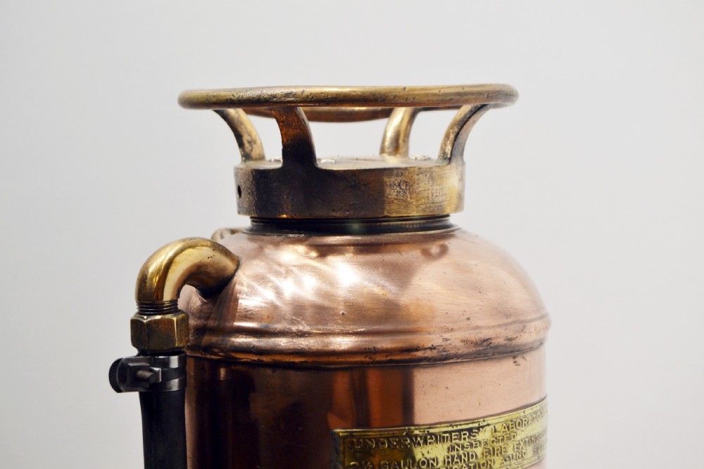 A cylindrical antique copper fire extinguisher featuring brass handles, brass instruction plaques, and a side-mounted black rubber hose. View II
