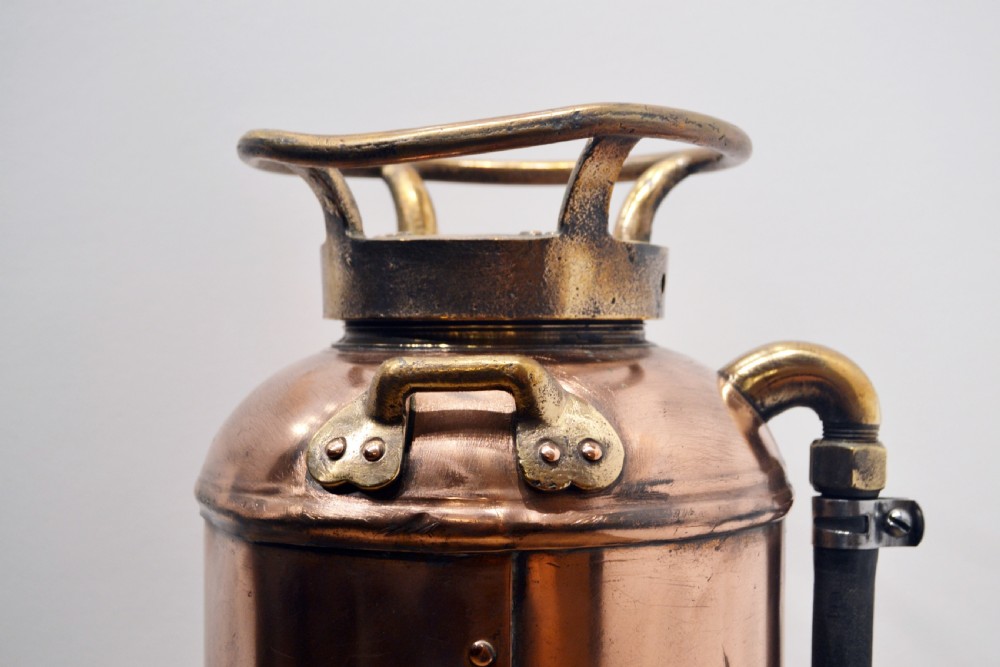 A cylindrical antique copper fire extinguisher featuring brass handles, brass instruction plaques, and a side-mounted black rubber hose. View V