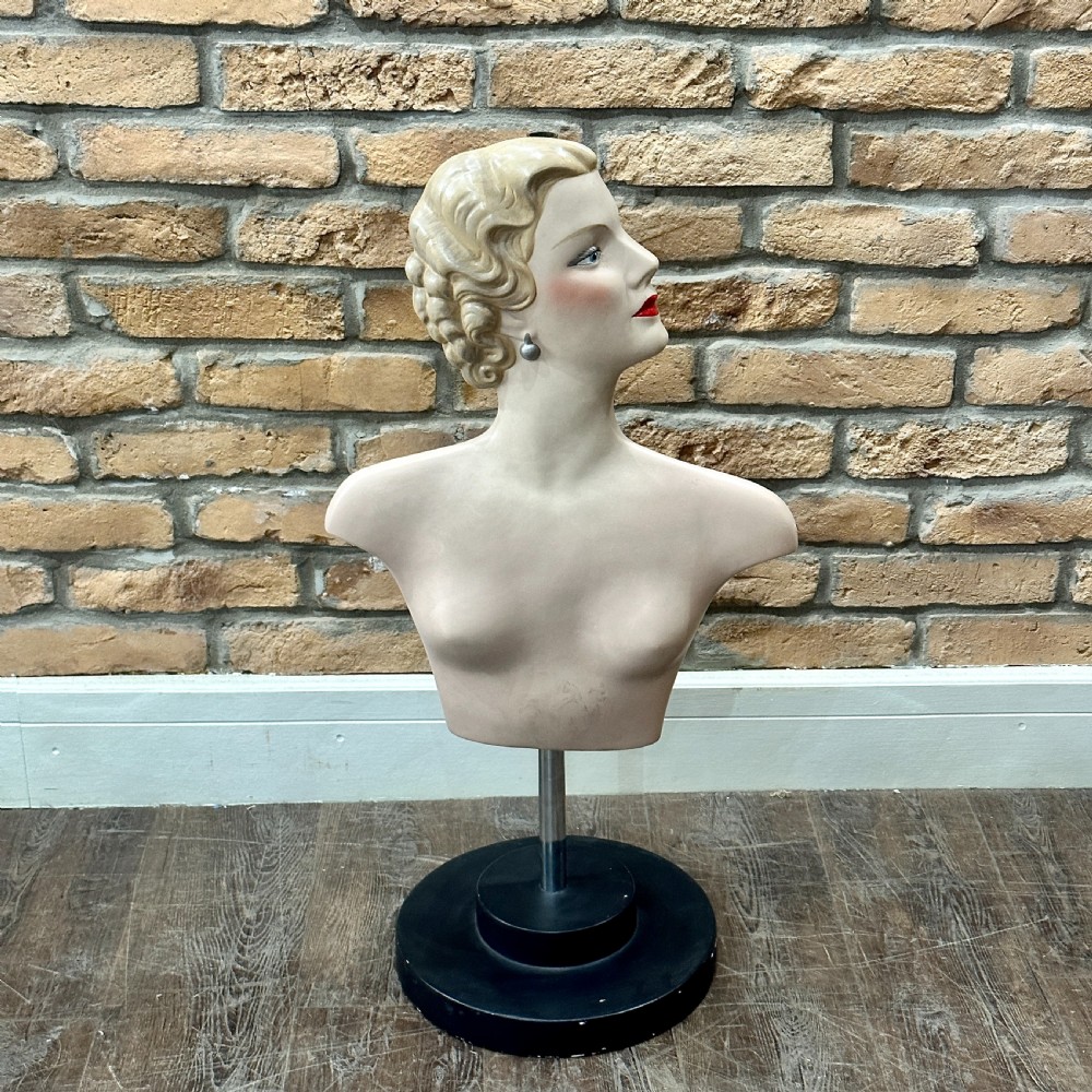 An Art Deco female mannequin bust featuring sculpted blonde waved hair, painted makeup, and a black circular stand.