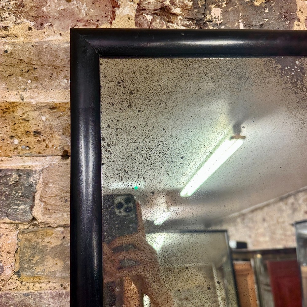 A tall rectangular full-length floor mirror with a slender black timber frame and foxed mercury glass plate. View III