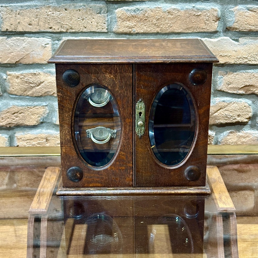 An Edwardian dark oak desktop smokers cabinet featuring twin doors with oval glass aperture windows and brass fittings.