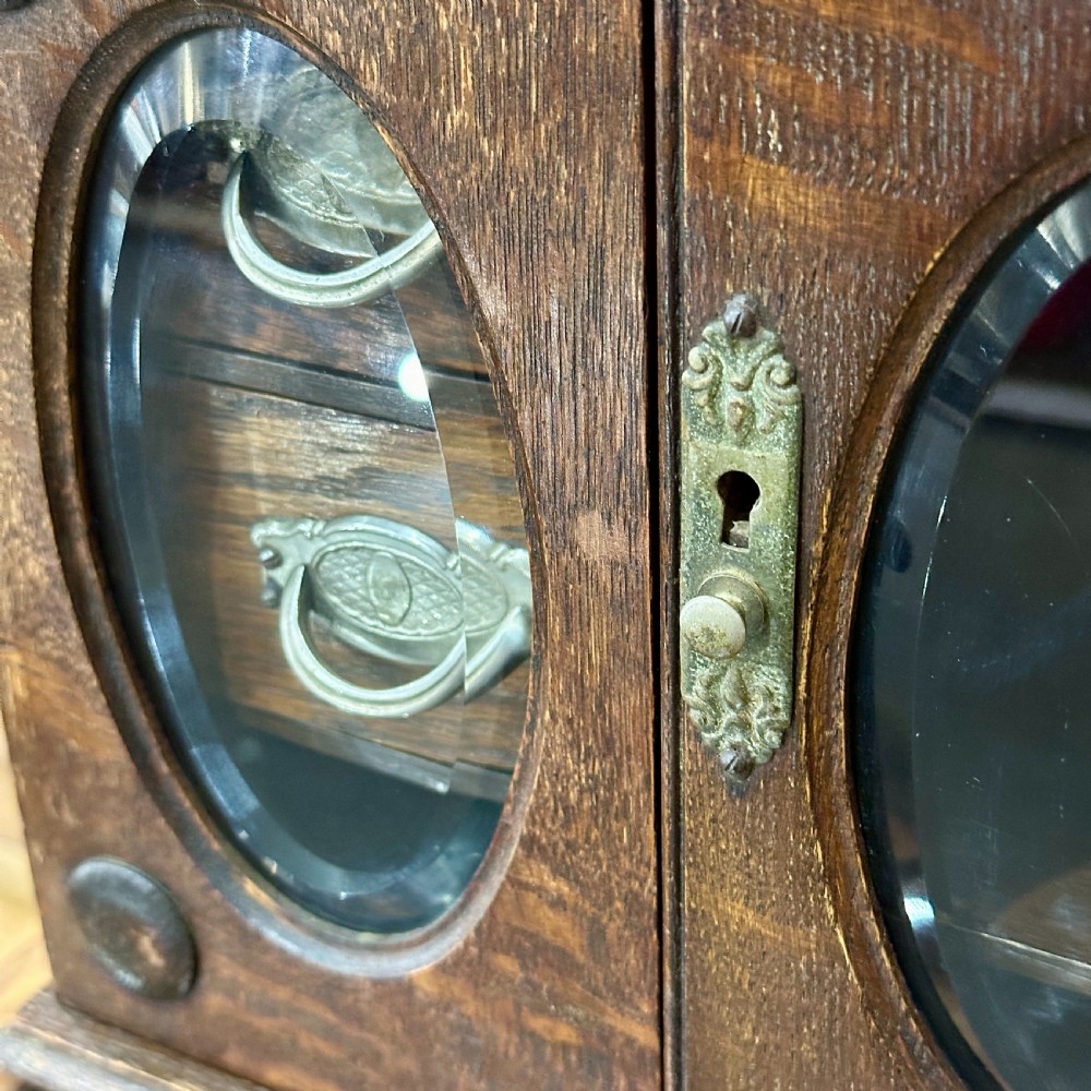 An Edwardian dark oak desktop smokers cabinet featuring twin doors with oval glass aperture windows and brass fittings. View II
