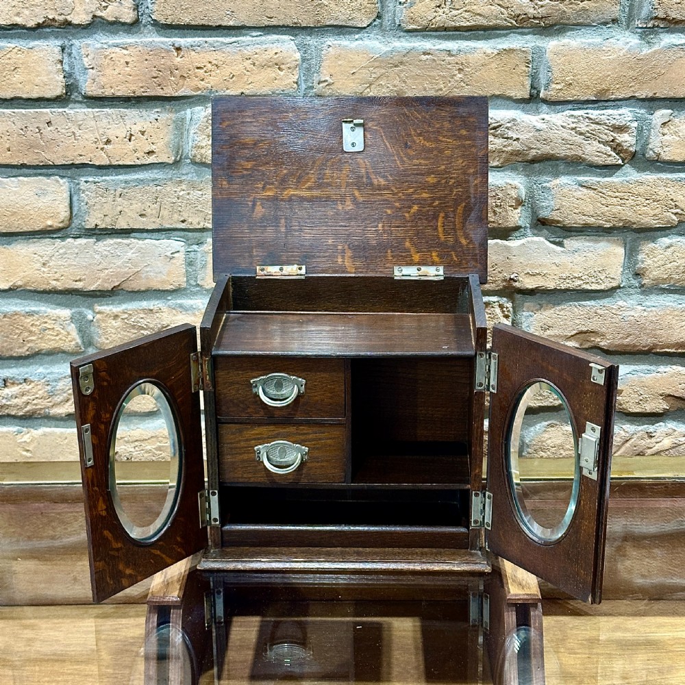 An Edwardian dark oak desktop smokers cabinet featuring twin doors with oval glass aperture windows and brass fittings. View III