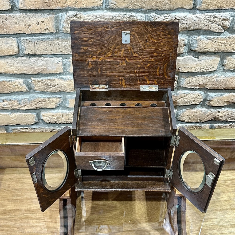 An Edwardian dark oak desktop smokers cabinet featuring twin doors with oval glass aperture windows and brass fittings. View IV