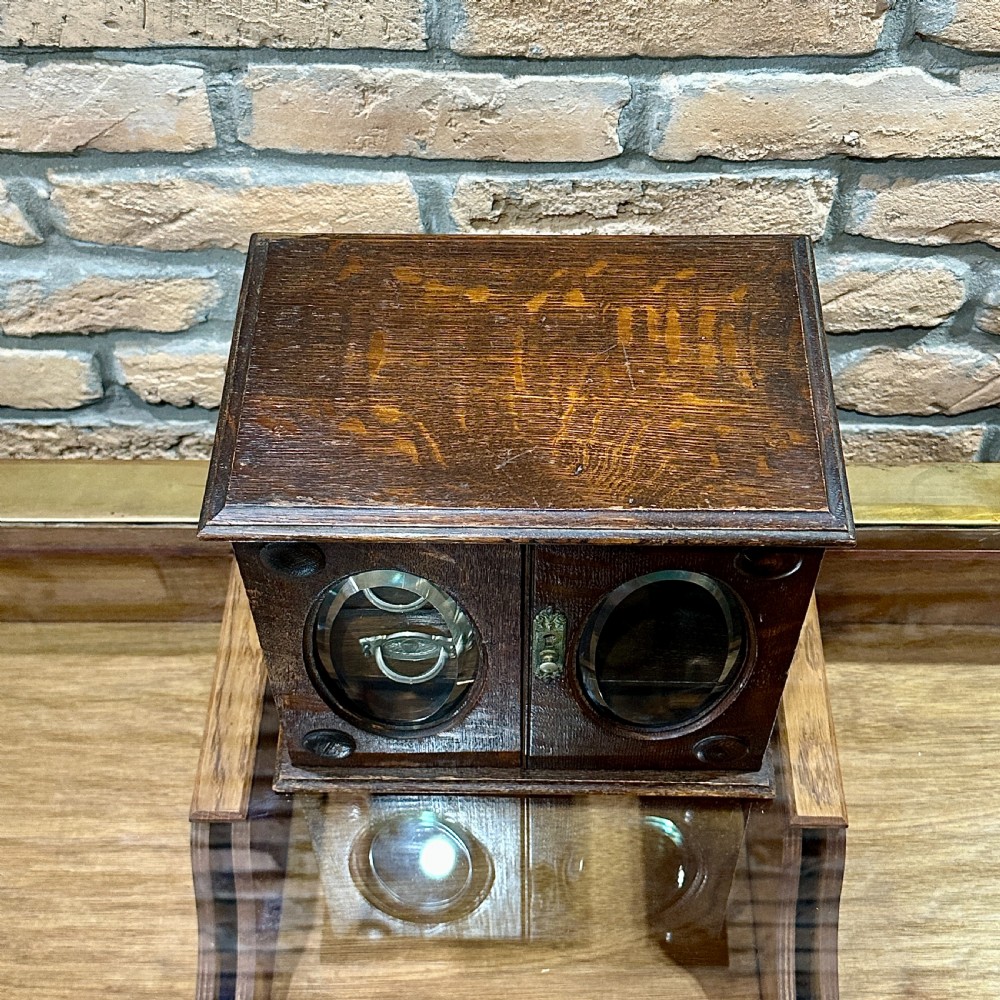 An Edwardian dark oak desktop smokers cabinet featuring twin doors with oval glass aperture windows and brass fittings. View V