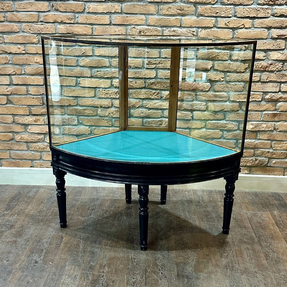 A 1920s quadrant-shaped ebonised shop display counter featuring all-around clear glass panels, turned legs, and a teal fabric-lined base.