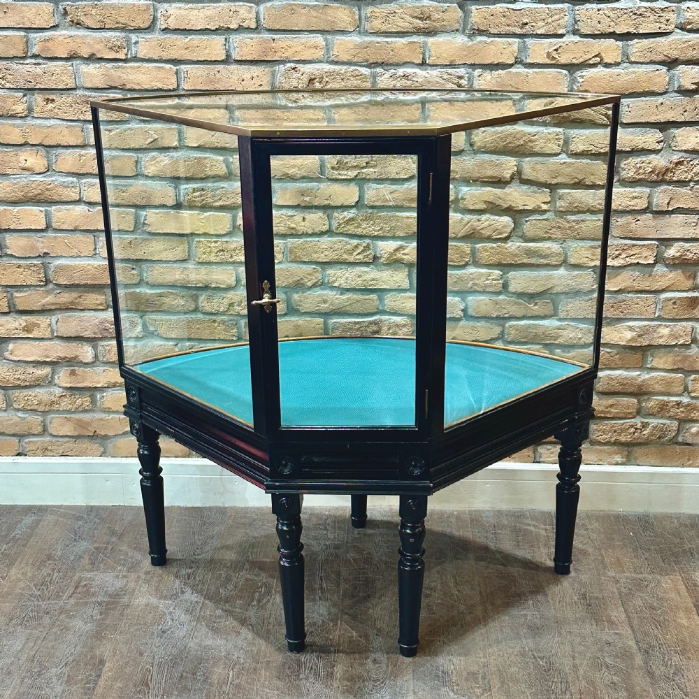 A 1920s quadrant-shaped ebonised shop display counter featuring all-around clear glass panels, turned legs, and a teal fabric-lined base. View II