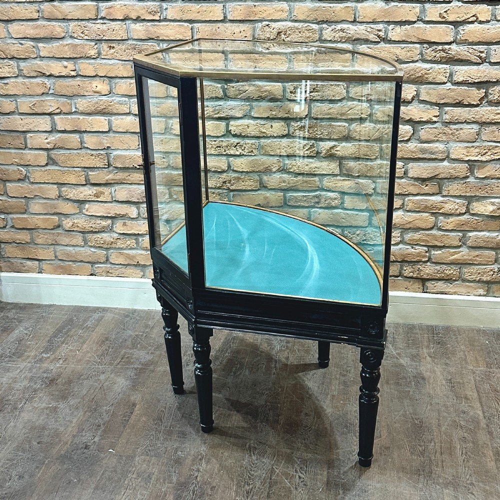 A 1920s quadrant-shaped ebonised shop display counter featuring all-around clear glass panels, turned legs, and a teal fabric-lined base. View III