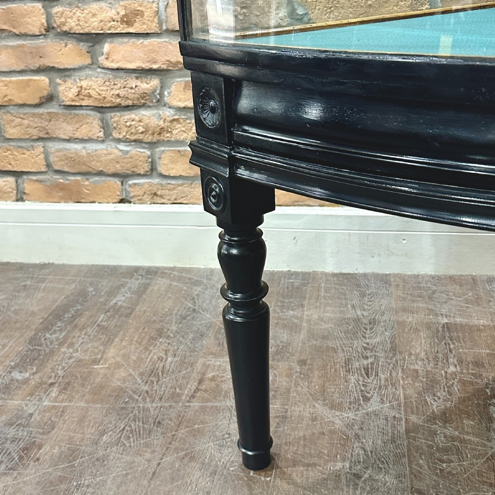 A 1920s quadrant-shaped ebonised shop display counter featuring all-around clear glass panels, turned legs, and a teal fabric-lined base. View IV