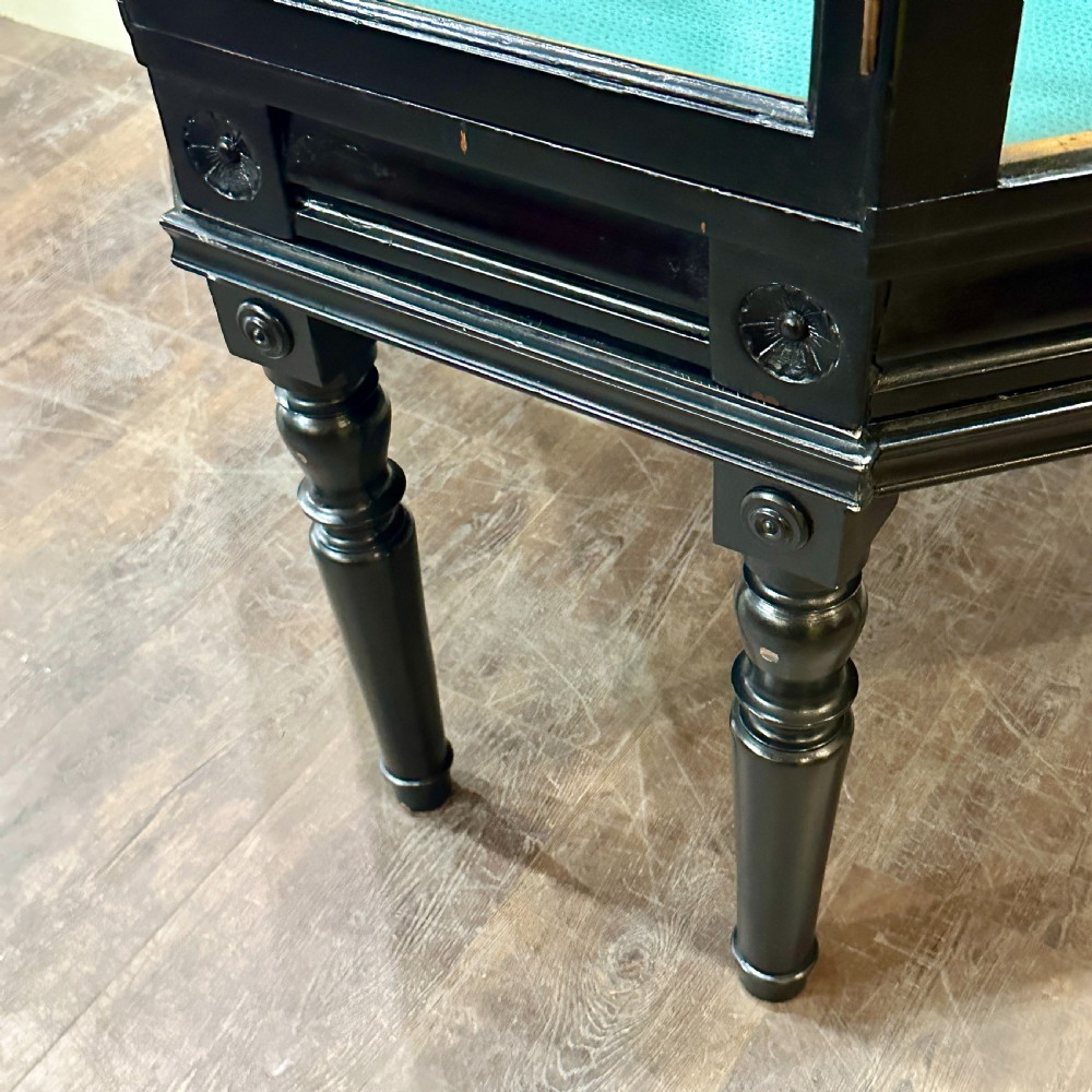 A 1920s quadrant-shaped ebonised shop display counter featuring all-around clear glass panels, turned legs, and a teal fabric-lined base. View VII