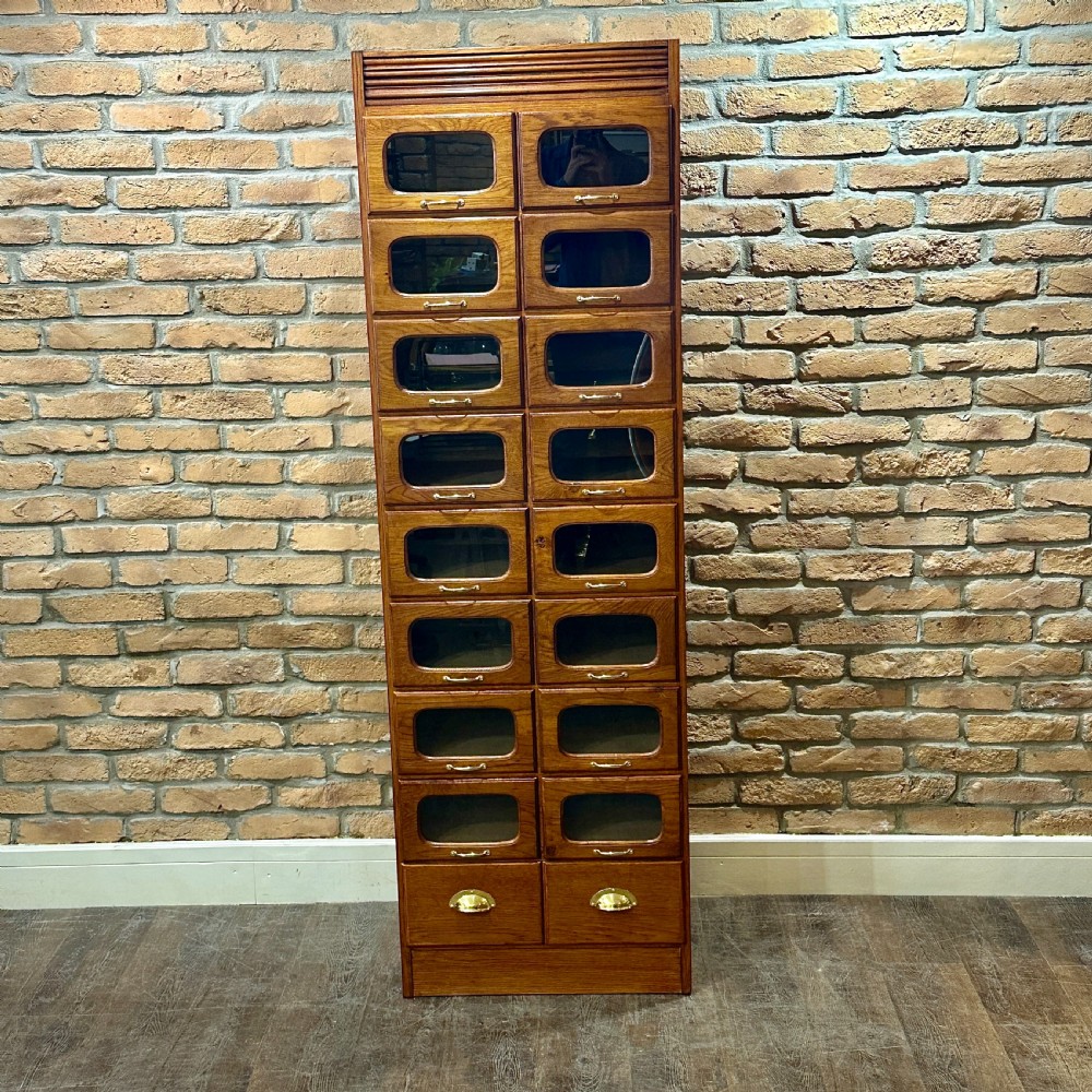 A tall, refurbished 1930s oak haberdashery cabinet featuring multiple glass-fronted drawers and brass hardware against a brick wall.