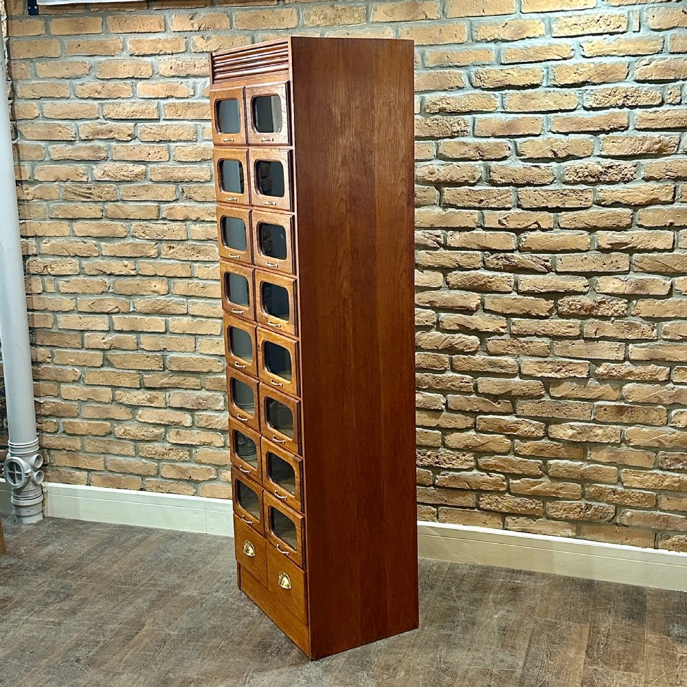 A tall, refurbished 1930s oak haberdashery cabinet featuring multiple glass-fronted drawers and brass hardware against a brick wall. View II