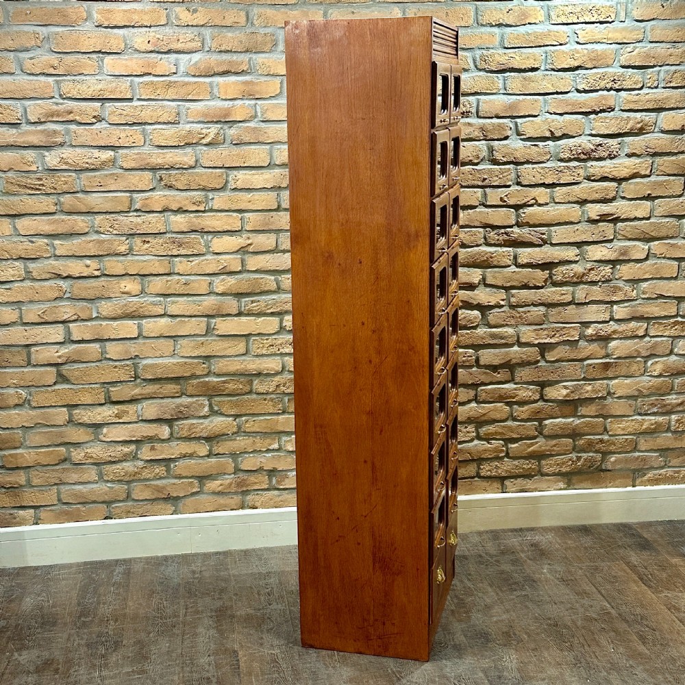 A tall, refurbished 1930s oak haberdashery cabinet featuring multiple glass-fronted drawers and brass hardware against a brick wall. View III