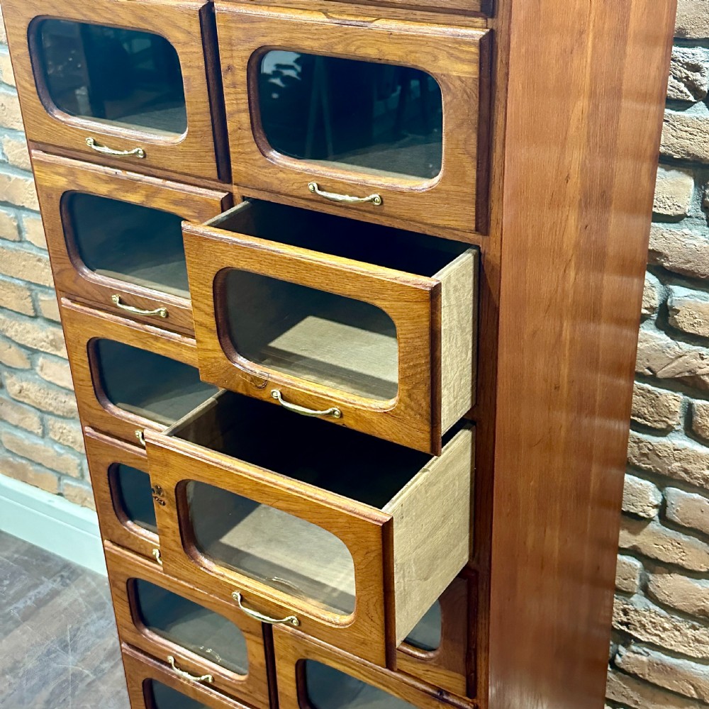 A tall, refurbished 1930s oak haberdashery cabinet featuring multiple glass-fronted drawers and brass hardware against a brick wall. View V