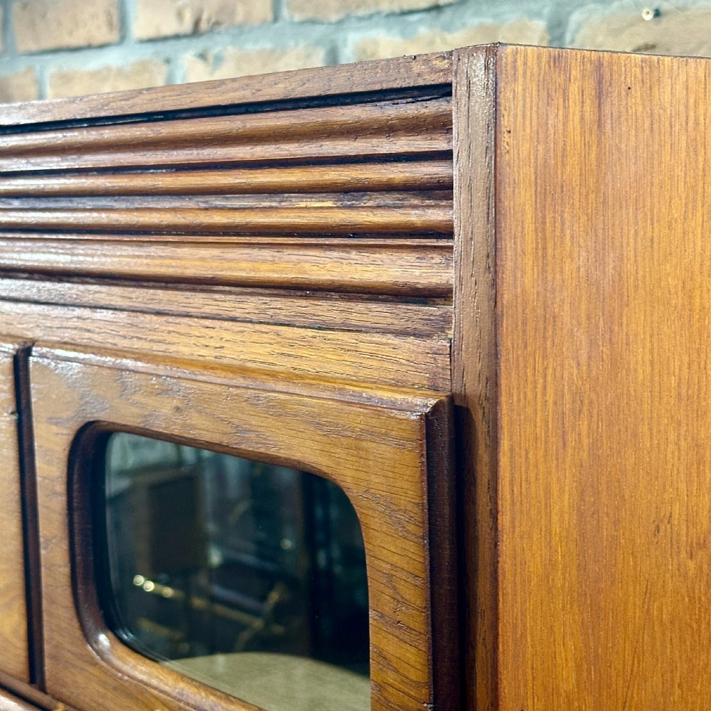 A tall, refurbished 1930s oak haberdashery cabinet featuring multiple glass-fronted drawers and brass hardware against a brick wall. View VI