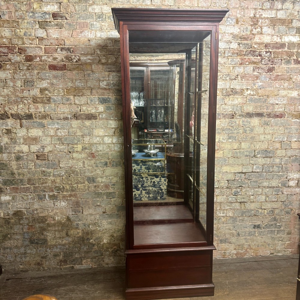 A tall 1920s mahogany shop display cabinet featuring full-length glass side panels, a glazed front door, internal shelf supports, and a solid timber plinth base.