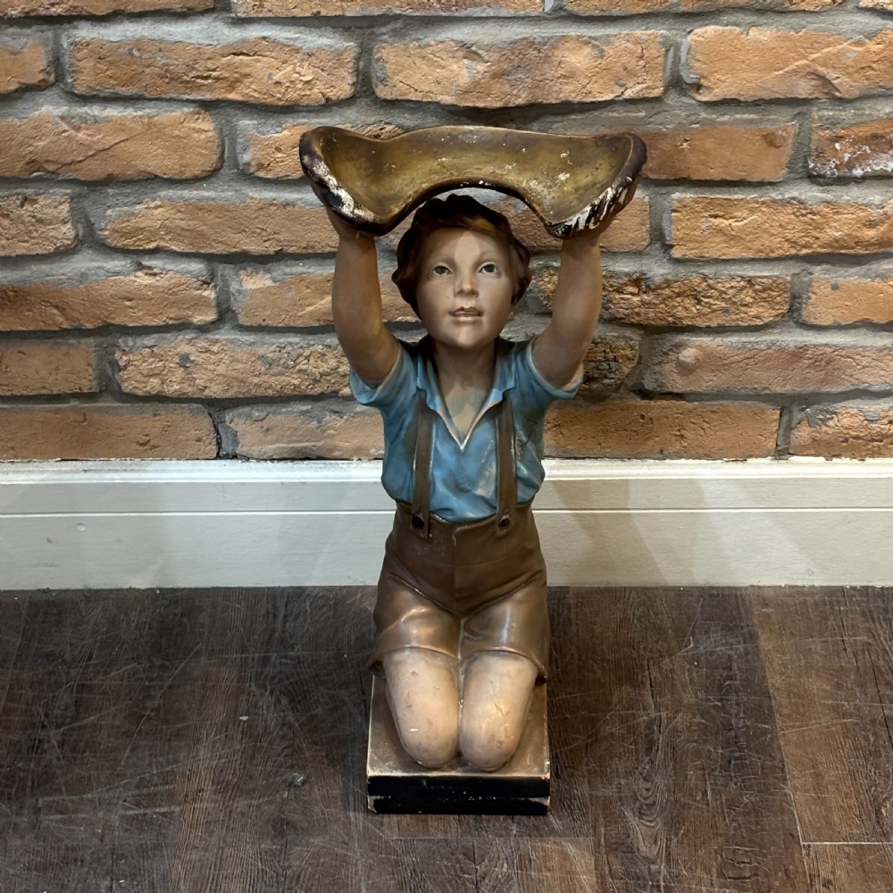 A hand-painted 1920s plaster sculpture of a kneeling boy wearing a blue shirt and brown shorts, supporting a shell-shaped tray overhead.