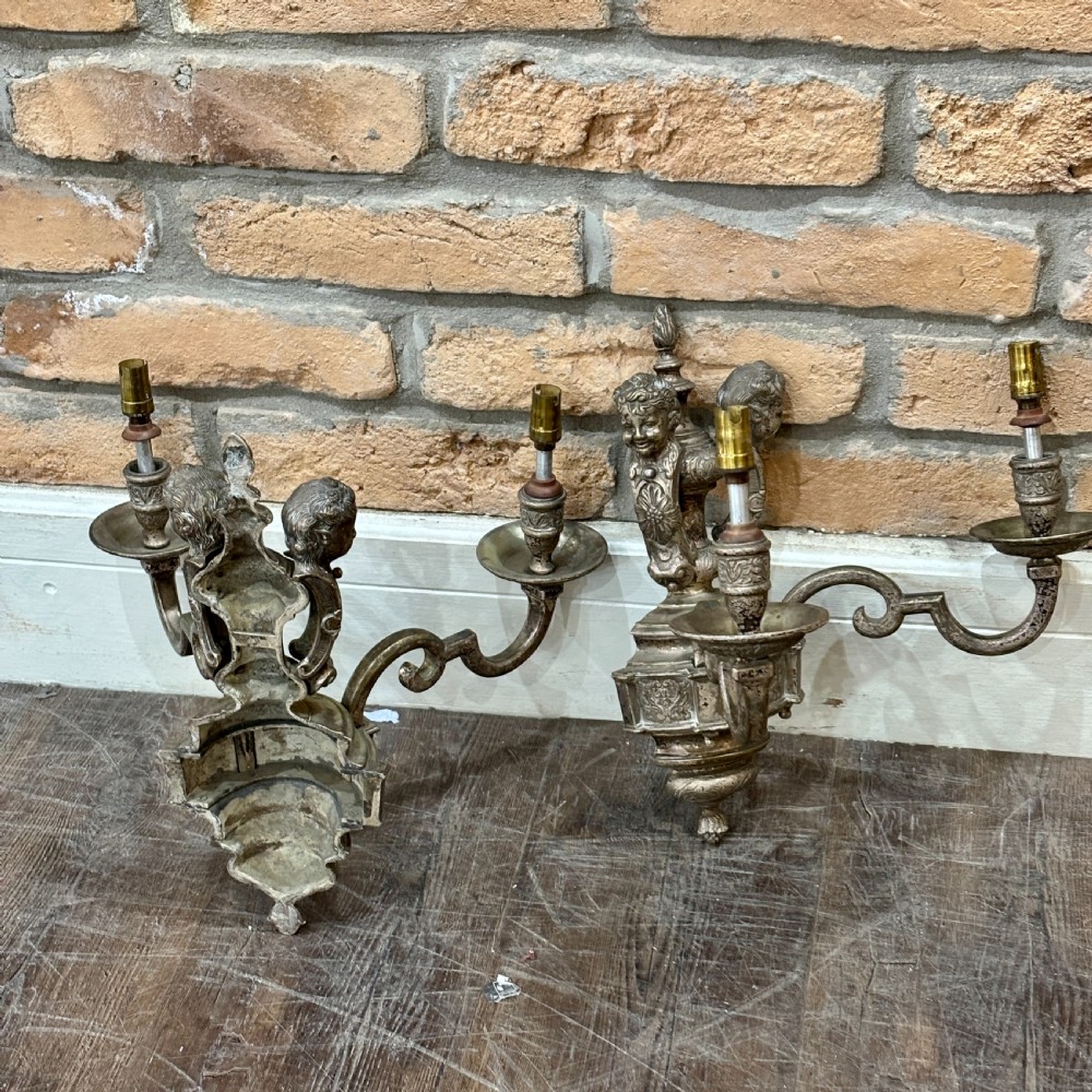 A pair of ornate cast brass twin-arm wall sconces featuring sculpted cherub heads and decorative scrolling branches.