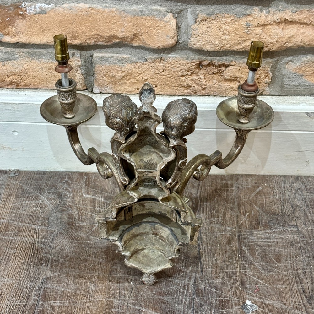 A pair of ornate cast brass twin-arm wall sconces featuring sculpted cherub heads and decorative scrolling branches. View VI