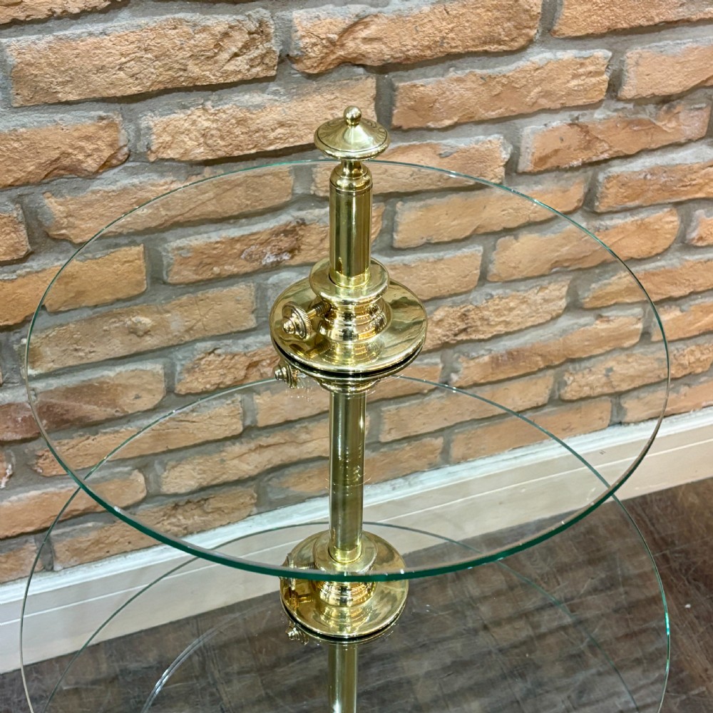 A three-tiered display stand featuring circular clear glass shelves mounted on a central polished brass column with a weighted circular base. View II