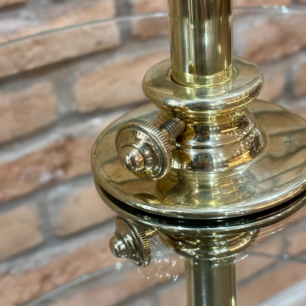A three-tiered display stand featuring circular clear glass shelves mounted on a central polished brass column with a weighted circular base. View VI