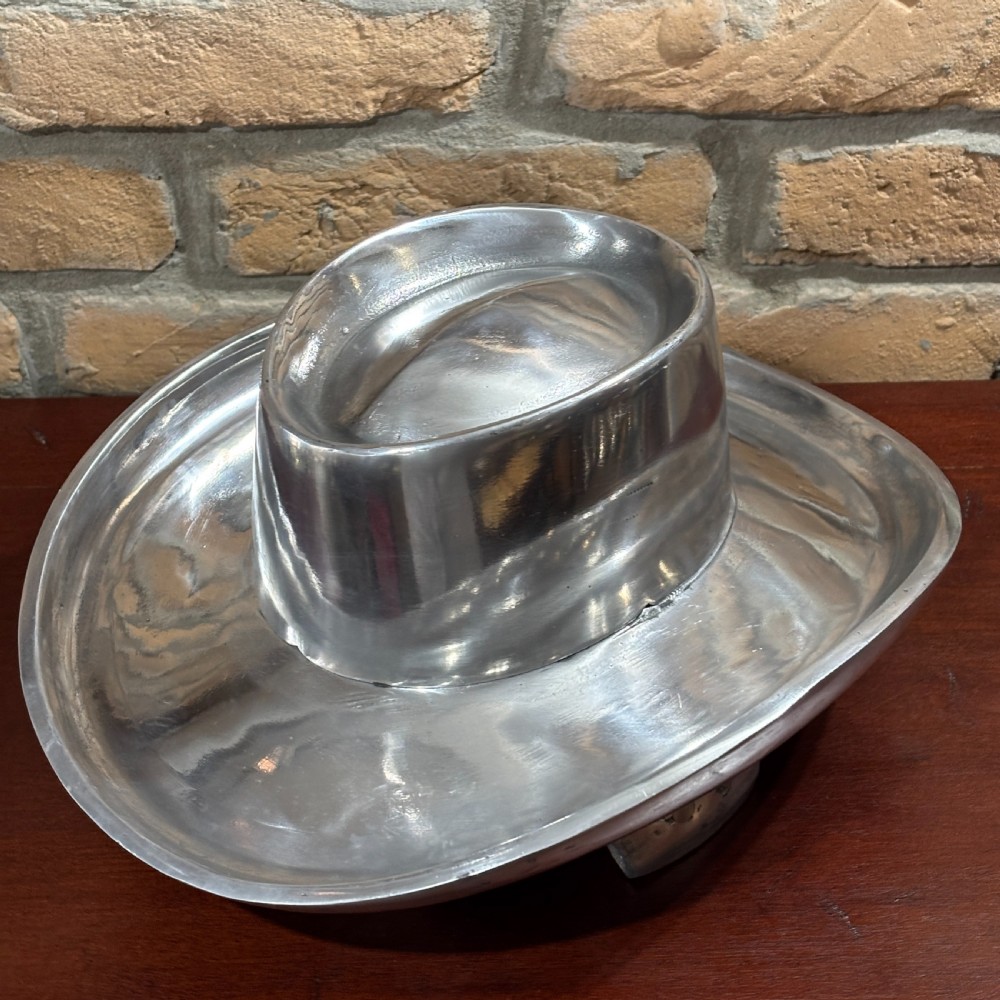 A cast aluminium milliners hat block mould in the shape of a wide-brimmed hat sitting on a polished dark wood surface. View III