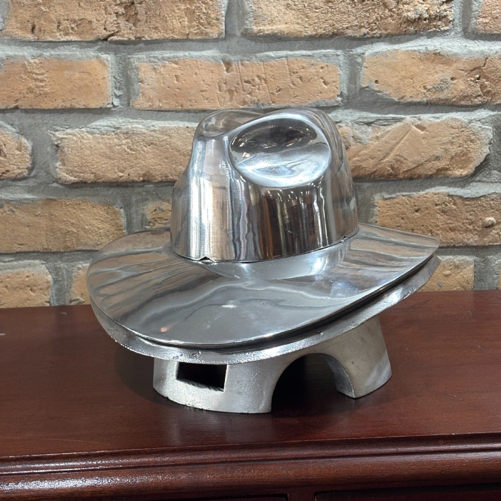 A polished silver aluminium milliners hat block mould shaped like a fedora resting on a wooden surface in front of a brick wall.